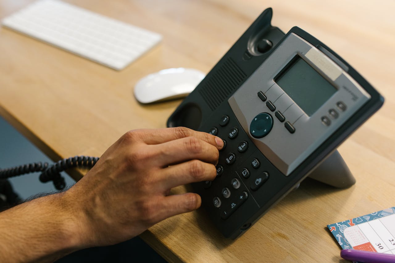 gallery-6 A hand dialing a phone on a wooden desk. Office environment with keyboard and planner.